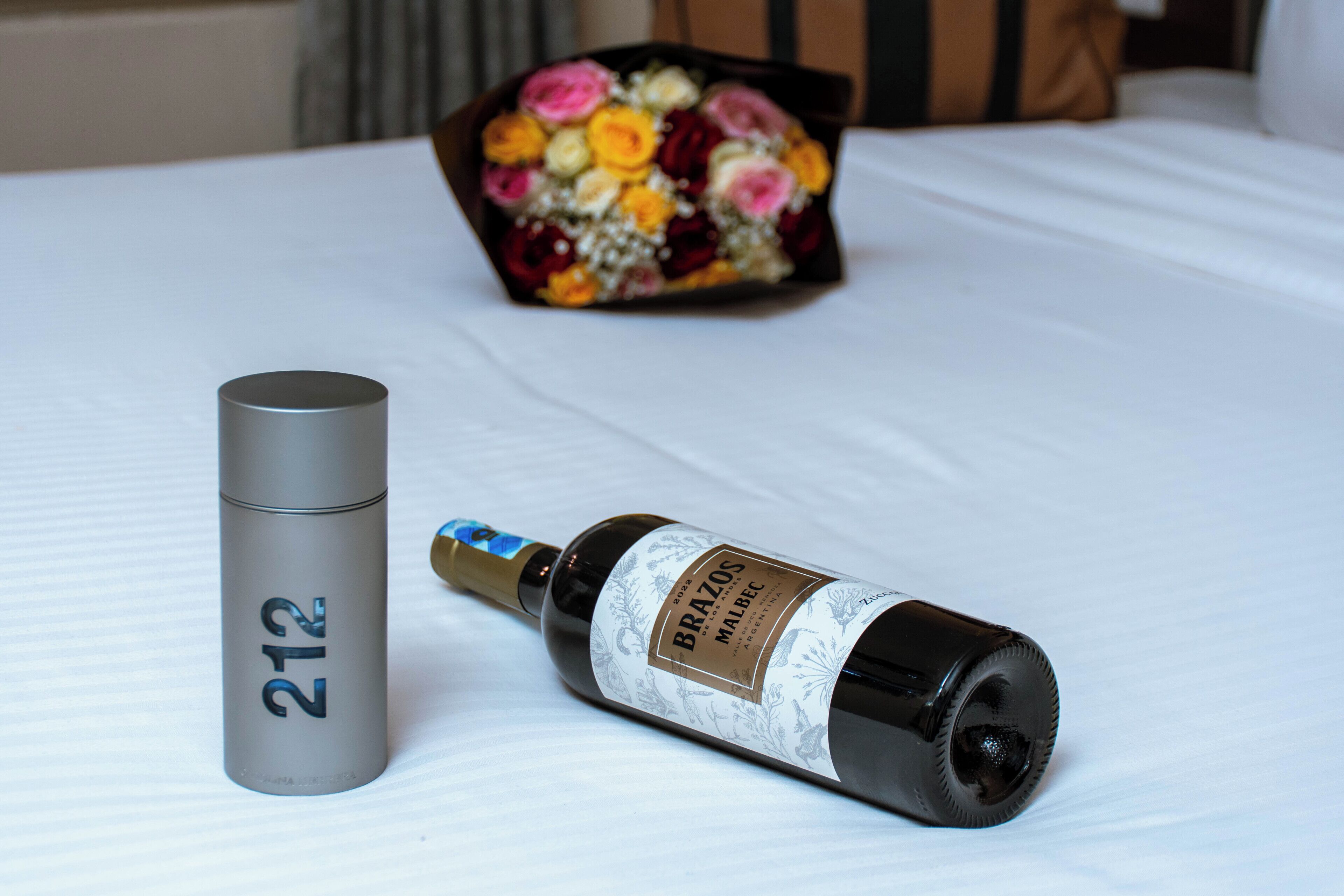 A bottle of wine, a cylindrical object, and a bouquet of flowers on a bed.