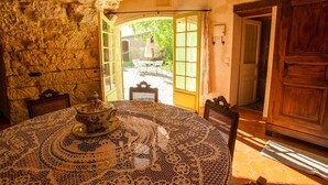 A stone-walled room with a round table covered by a lace tablecloth, a wooden cabinet, and a view of an outdoor garden through an open door.