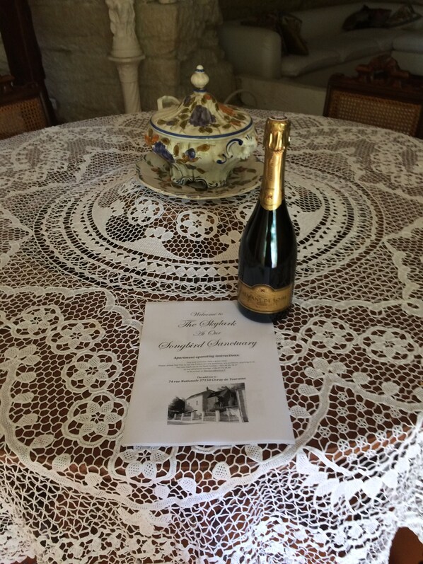 A table with a lace tablecloth, a bottle of champagne, and a cake on a stand. There is a welcome card with the address and operating instructions.