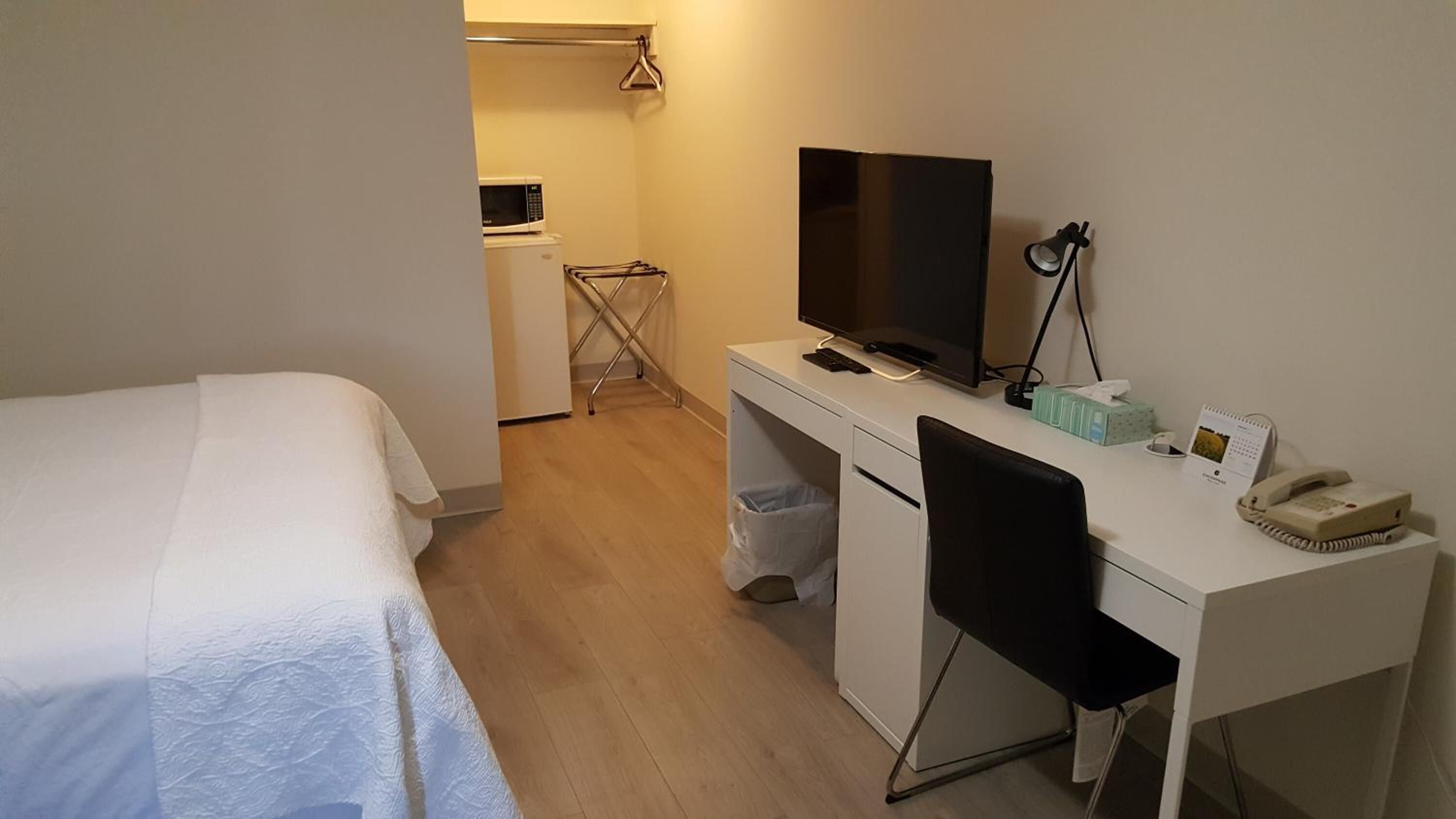 A hotel room with a bed, a desk with a TV, a chair, a trash bin, a lamp, a telephone, and a calendar.