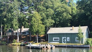 Ein an einem Steg festgemachtes Boot nahe einem grünen Haus mit weißer Tür und Fenstern, umgeben von Bäumen und einem Gewässer.