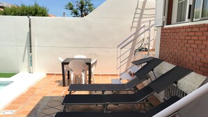 A sunlit outdoor seating area with lounge chairs and a table, adjacent to a swimming pool and a brick wall. - Private pool beach. Sohail Castle (Fuengirola)