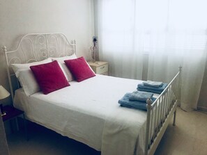 Ein ordentlich bezogenes Bett mit weißer Bettwäsche und zwei roten Kissen, ein Nachttisch mit Lampe sowie ein Fenster mit durchsichtigen Vorhängen.