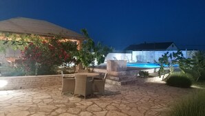 Un espace extérieur aménagé en terrasse, avec un sol en pierre, une piscine et un coin salon couvert avec des chaises.