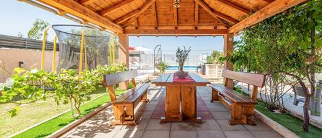 A wooden pergola with a table and benches, surrounded by greenery and a clear view of the sky.