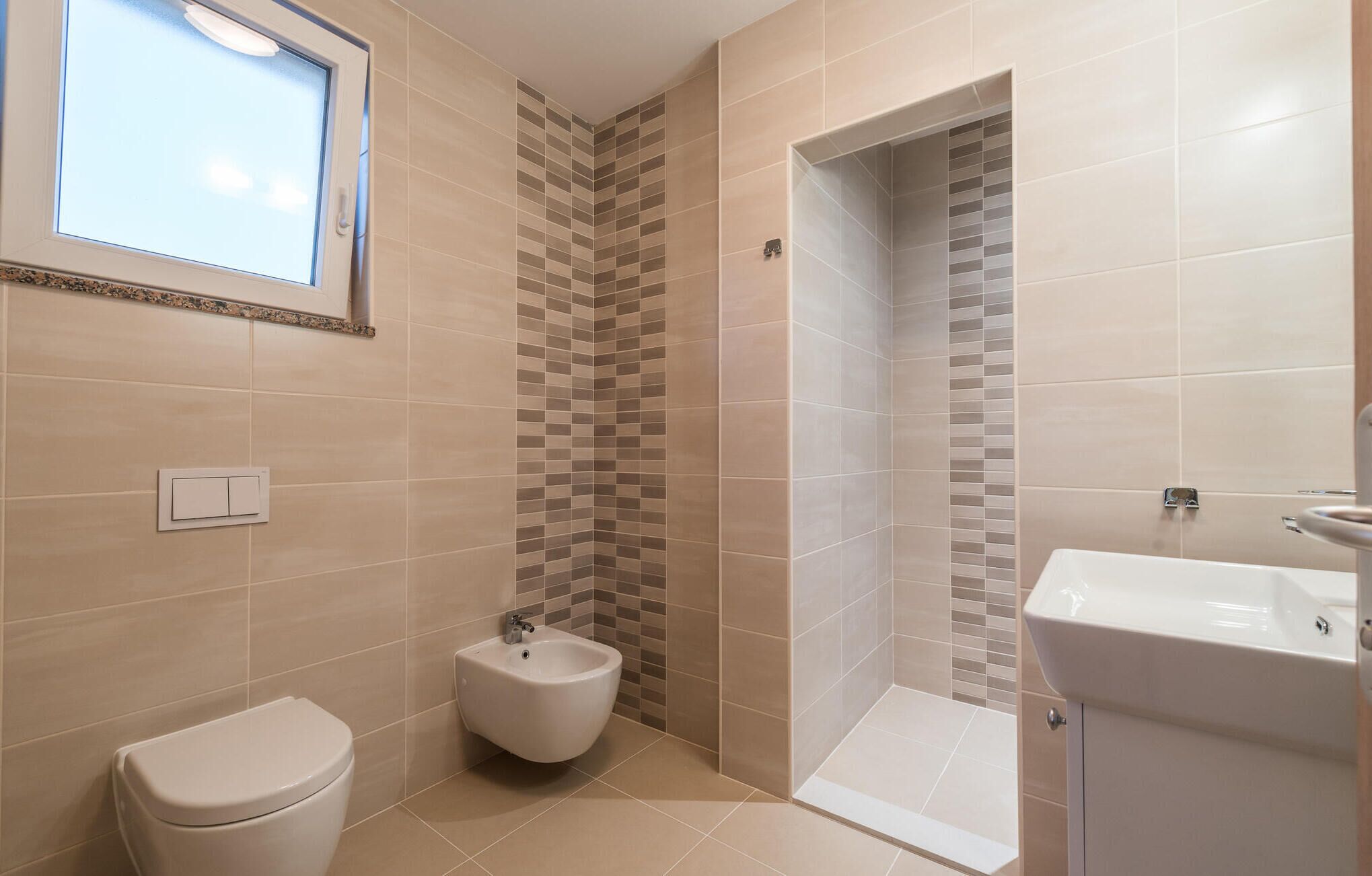 Ein Badezimmer mit Toilette, Bidet und Waschbecken, ausgestattet mit beigen Fliesen und einem Fenster.