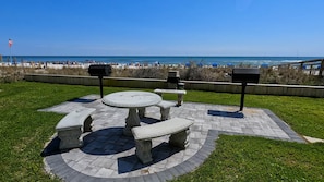 Un patio con una mesa redonda y bancos, rodeado de baldosas de piedra y césped, con vistas a una playa donde hay gente y barcos.