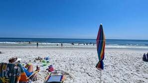 Una playa con gente nadando y jugando, una sombrilla colorida y una silla de playa con una toalla.