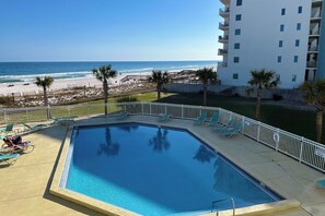 A swimming pool with a view of the beach and ocean, a fenced area with lounge chairs, and a multi-story building in the background.