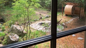 Vista a través de una ventana que muestra una zona boscosa con un sendero de piedra, barriles de madera y un pequeño árbol.