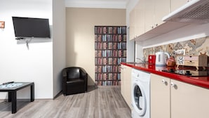 Ein gemütlicher Wohnbereich mit Flachbildfernseher, Bücherregal, schwarzem Ledersessel, Waschmaschine und roter Arbeitsplatte mit Küchengeräten.