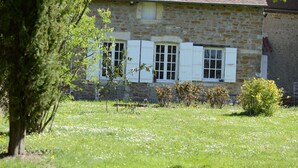 Un edificio de piedra con contraventanas blancas, rodeado por un campo cubierto de pasto y flores blancas.