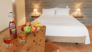 Een opgemaakt bed met witte lakens, een houten nachtkastje met een lamp, en een tafel met fruit en een fles drank.
