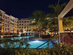 A nighttime view of a hotel pool area with a large pool, palm trees, and a multi-story building in the background.