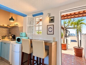A compact kitchen with a coffee machine, a toaster, and a refrigerator. There is a dining area with two chairs and a table. A window with frosted glass and a picture frame are visible. - Le Petit Bonheur • Quiet • Rooftop • 3 min to Beach (5 de Diciembre)