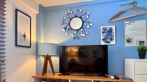 A bedroom with a TV on a wooden stand, a bedside table with a lamp, a mirror, and a wall-mounted drying rack. - Le Petit Bonheur • Quiet • Rooftop • 3 min to Beach (5 de Diciembre)