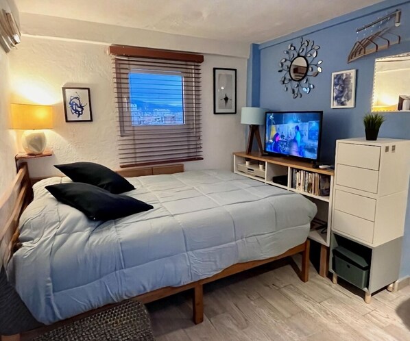 Une chambre à coucher comprenant un lit, une télévision, une fenêtre avec des stores, un miroir et une armoire.