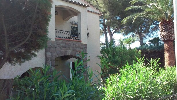 Un edificio con un arco de piedra, un balcón con baranda de metal y abundante vegetación alrededor.