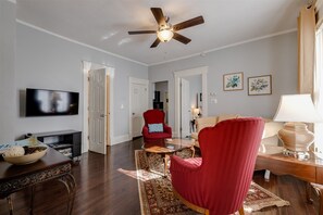 Una sala de estar con ventilador de techo, televisor, un sillón rojo, una mesa de centro de madera y un sofá beige.