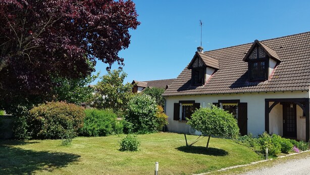 Ein traditionelles Haus mit einem geneigten Dach, einem Schornstein und einem kleinen Fenster. Vor dem Haus erstreckt sich ein gepflegter Rasen und ein Kiesweg führt zum Haus.