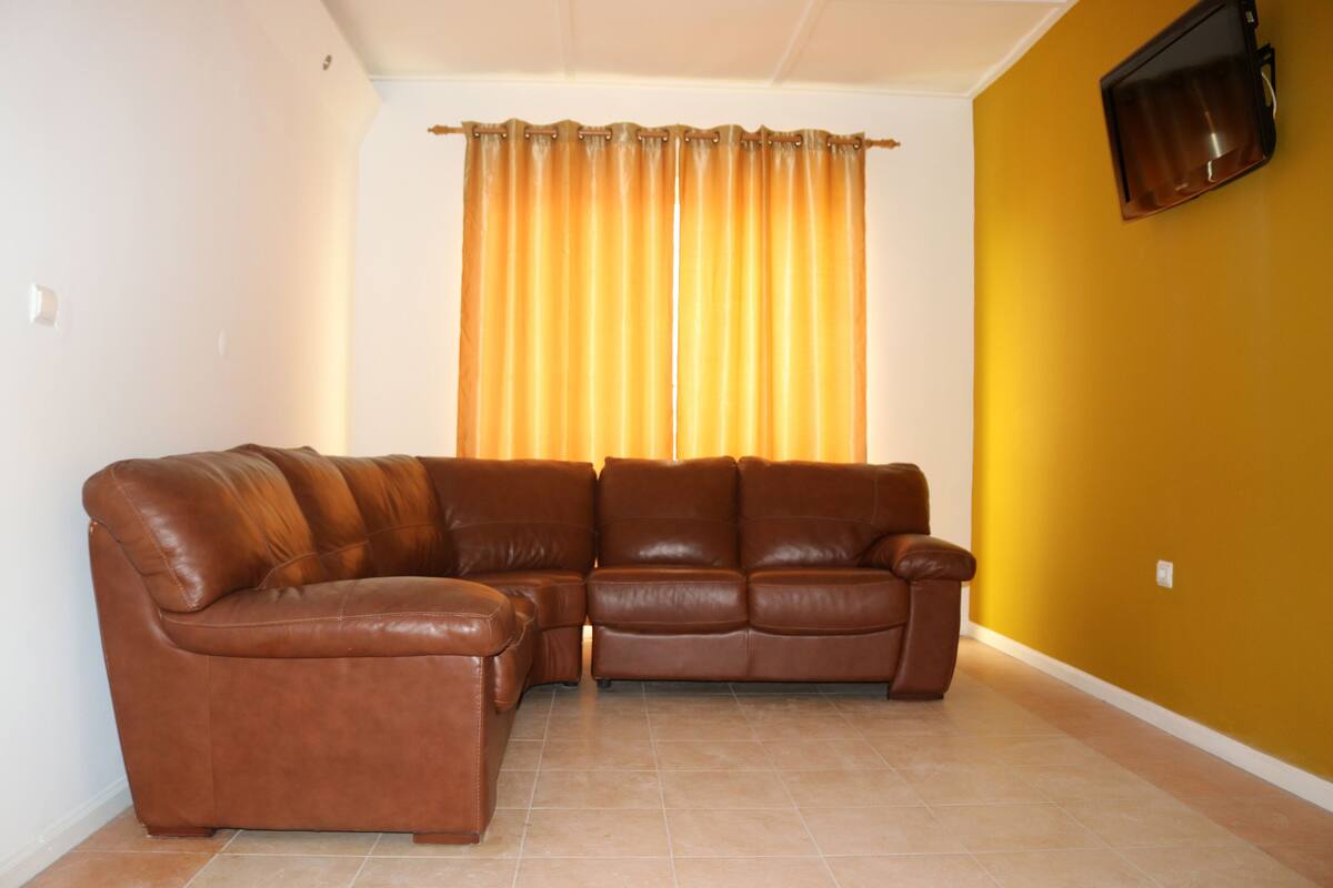 A living room with a brown leather sofa, a yellow wall, and a window with curtains.
