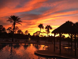 Zwembadgebied van een resort met uitzicht op een zonsondergang, palmbomen en een constructie met rieten dak.