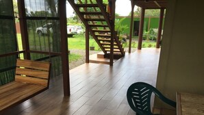 Un espacio exterior cubierto con un banco de madera, una silla de plástico verde y una escalera que conduce a un nivel superior.