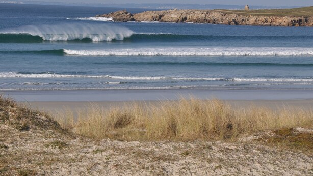Uma praia costeira com ondas, um afloramento rochoso e uma área gramada.