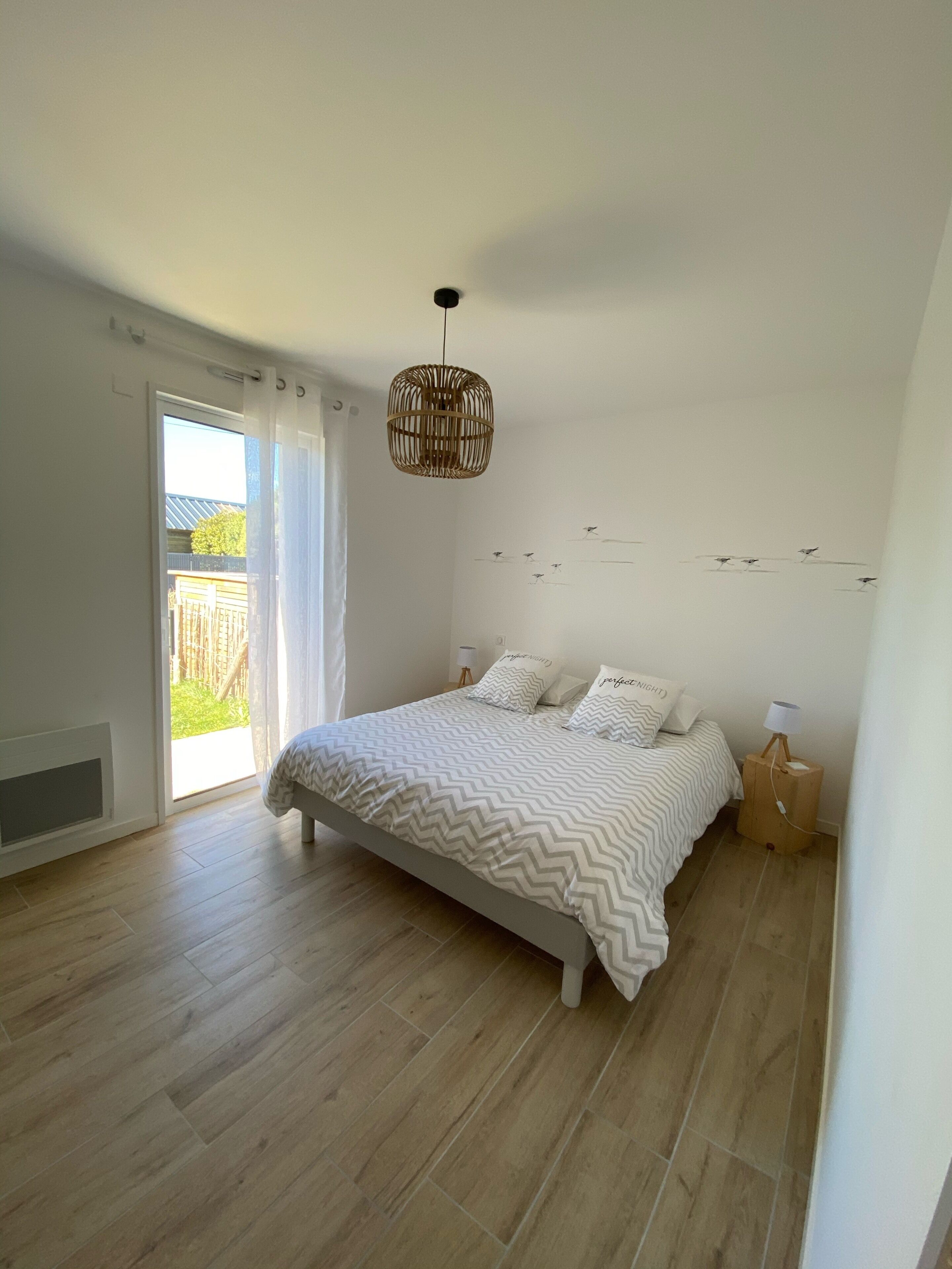 A bedroom with a bed, a hanging light fixture, a nightstand, and a view of the outdoors.