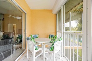 A balcony with a white table and chairs, a glass door leading to a room, and a view of greenery outside. - Resort-style condo - close to great beaches and downtown Naples. (Naples)