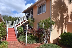 A two-story house with a red brick staircase, a balcony, and a variety of tropical plants.