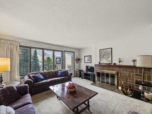 A living room with a sofa, coffee table, fireplace, and large windows offering a view of trees and hills. - 2 bedroom Condo overlooking Lake Dillon CC43 (Dillon)