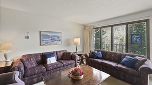 Ein Wohnzimmer mit zwei gemusterten Sofas, einem Holztisch mit Blumenarrangements, einer Lampe und einem großen Fenster mit Blick auf Bäume und einen Balkon.