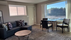 Un salon avec un canapé bleu foncé, deux fauteuils en osier noirs, une table basse ronde en bois et une grande fenêtre donnant sur une forêt et un lac.