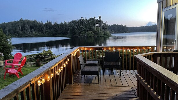 Une terrasse en bois, agrémentée de chaises et de guirlandes lumineuses, qui offre une vue sur un lac paisible et une zone boisée.