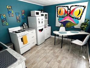 Een moderne keuken met een fornuis, magnetron en koelkast, een eethoek met een tafel en stoelen, en abstracte wanddecoratie.