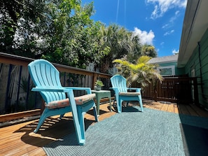 Een houten terras met twee blauwe Adirondack-stoelen en een kleine tafel, omgeven door weelderige begroeiing en een houten hek.