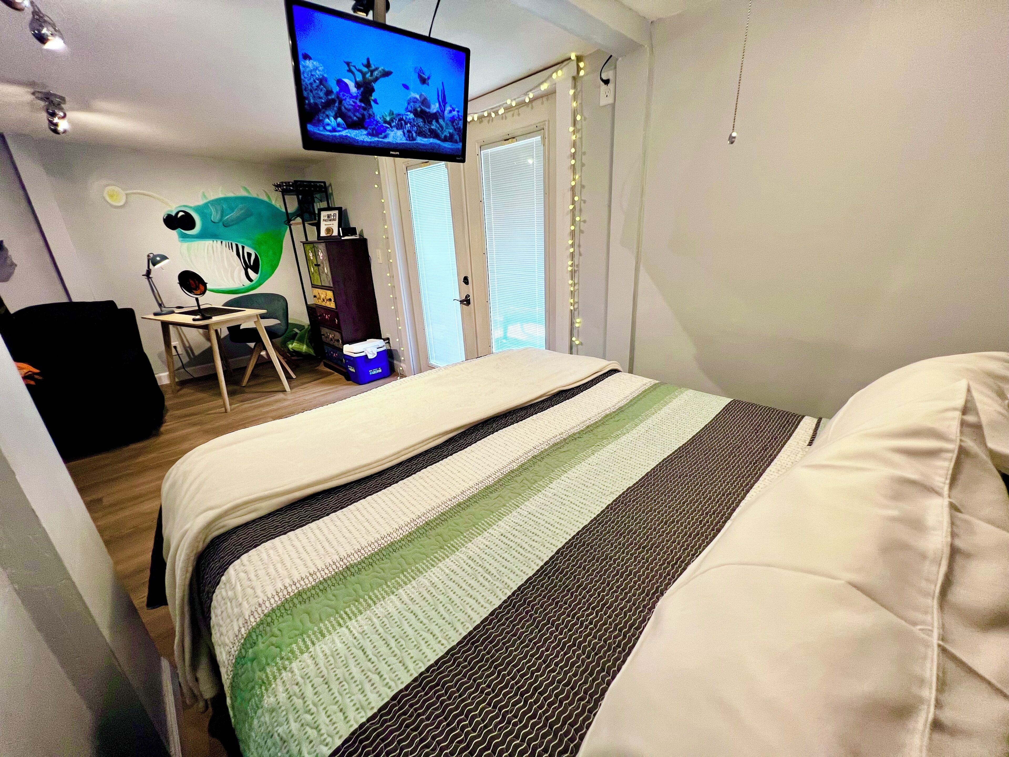 Ein Schlafzimmer mit einem Bett, einem Schreibtisch mit Computer, einem Fernseher und einer Tür mit Beleuchtung.