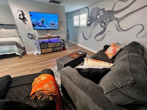Un salon comprenant un canapé, un téléviseur, une fresque murale, un lit et un meuble en bois.