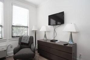 A room with a single chair, a dresser, a television mounted on the wall, and two lamps on either side of the dresser.