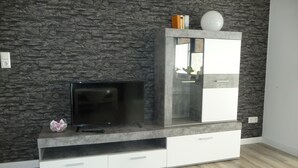 Un soggiorno moderno con una parete effetto pietra, una TV a schermo piatto su una mensola a muro e un mobile vetrina con finitura bianca.