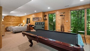Um quarto aconchegante com mesa de bilhar, cama com roupa de cama xadrez e uma grande janela com vista para a floresta.