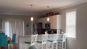 Una cocina moderna con armarios blancos, un microondas y un ventilador de techo. Hay un comedor con sillas blancas y un sofá verde azulado.