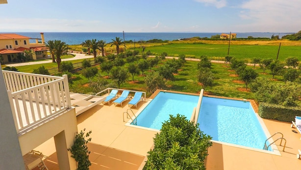A swimming pool with two lounge chairs, a view of the sea, and a grassy area with trees. - Betty's Villas (Pyrgos)
