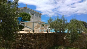 A stone wall with a white building on top, a blue chair, and a tree. - Betty's Villas (Pyrgos)