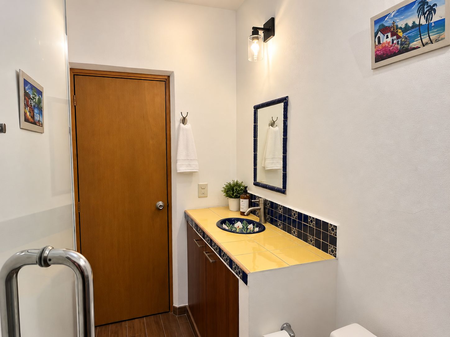 A bathroom with a wooden door, a sink with a blue-tiled backsplash, a mirror, and a wall-mounted light fixture.