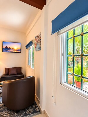 Ein Wohnzimmer mit einer Couch, einem Fernseher, der einen Sonnenuntergang zeigt, und einem Fenster mit Blick auf Grünflächen.