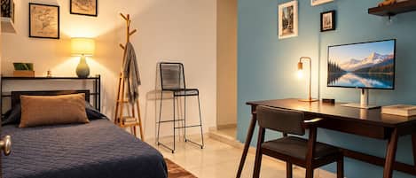 Un dormitorio con una cama, un escritorio con computadora, una silla, una lámpara y un perchero.