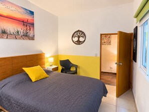 Ein Schlafzimmer mit einem Bett, einem Nachttisch, einem Sessel, einer Vogeldarstellung an der Wand, einer Baumdekoration und einer Tür zu einem anderen Raum.