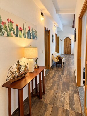 Un couloir avec un plancher en bois, une table avec une lampe, une chaise et un mur orné d’un tableau représentant un cactus.